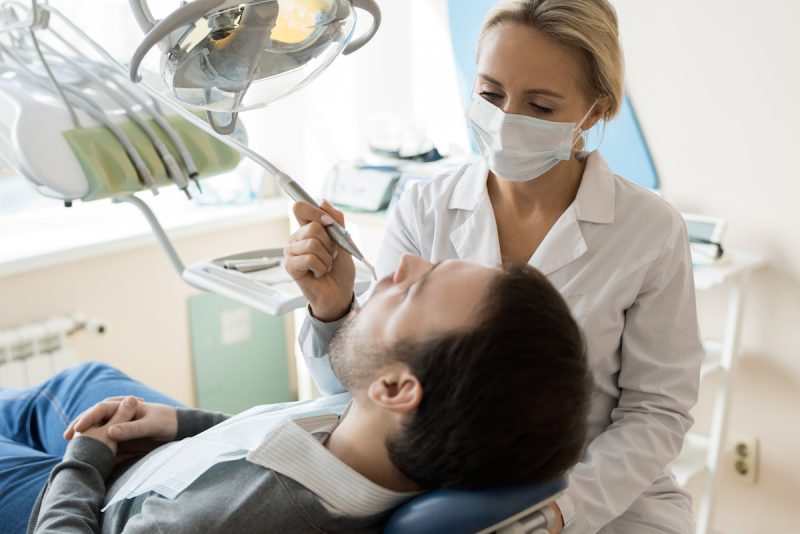 Portrait of blonde young woman treating teeth of relaxed man sitting in dental chair in modern clinic